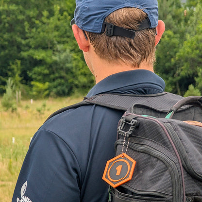 A man viewed from behind wearing a blue cap, navy shirt, and a black backpack with an orange hexagonal tag featuring the number 1.