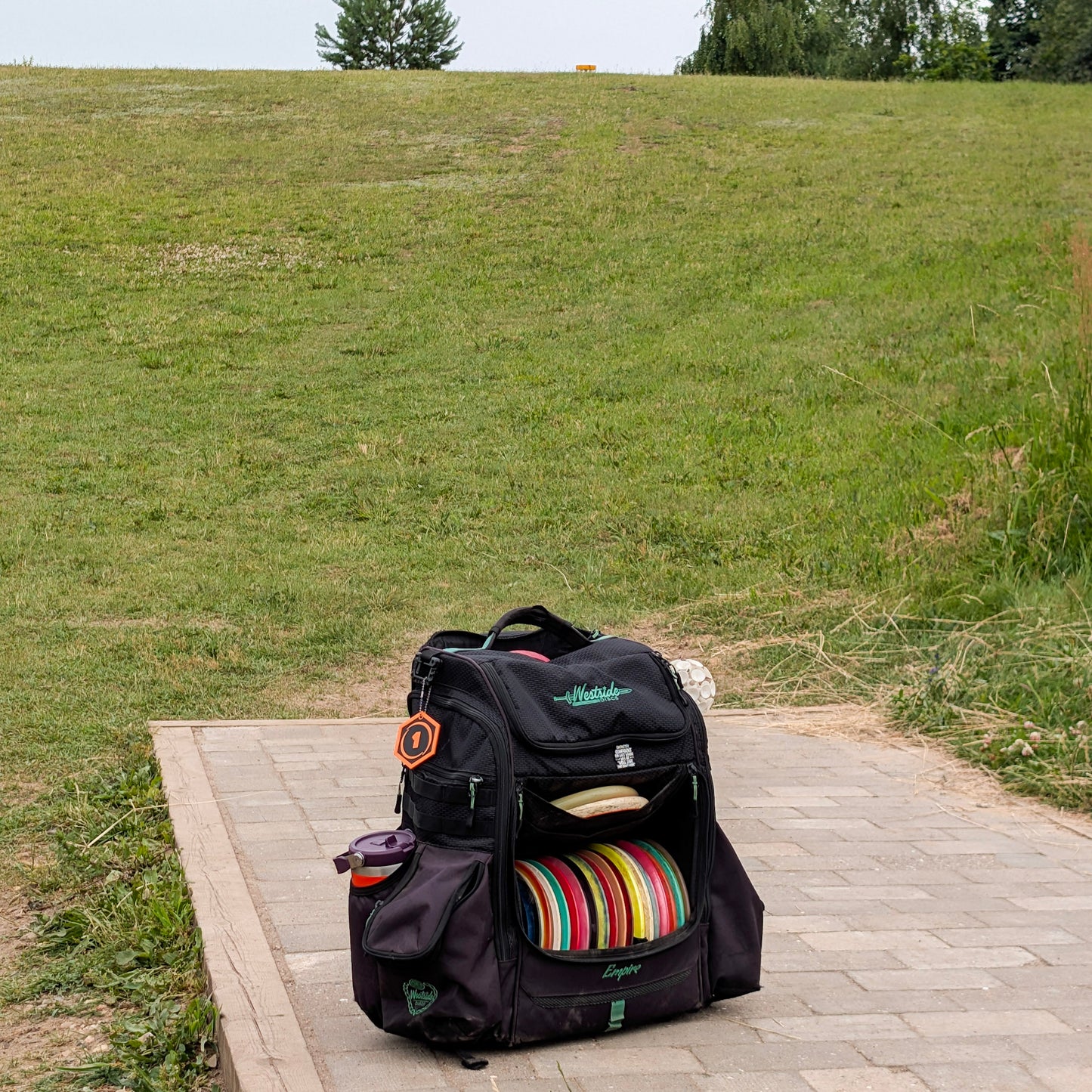 A black disc golf backpack sitting on a paved platform in an outdoor grassy field. The backpack has multiple compartments, a hexagonal disc golf bag tag, a side water bottle pocket, and a stack of colorful disc golf discs inside.