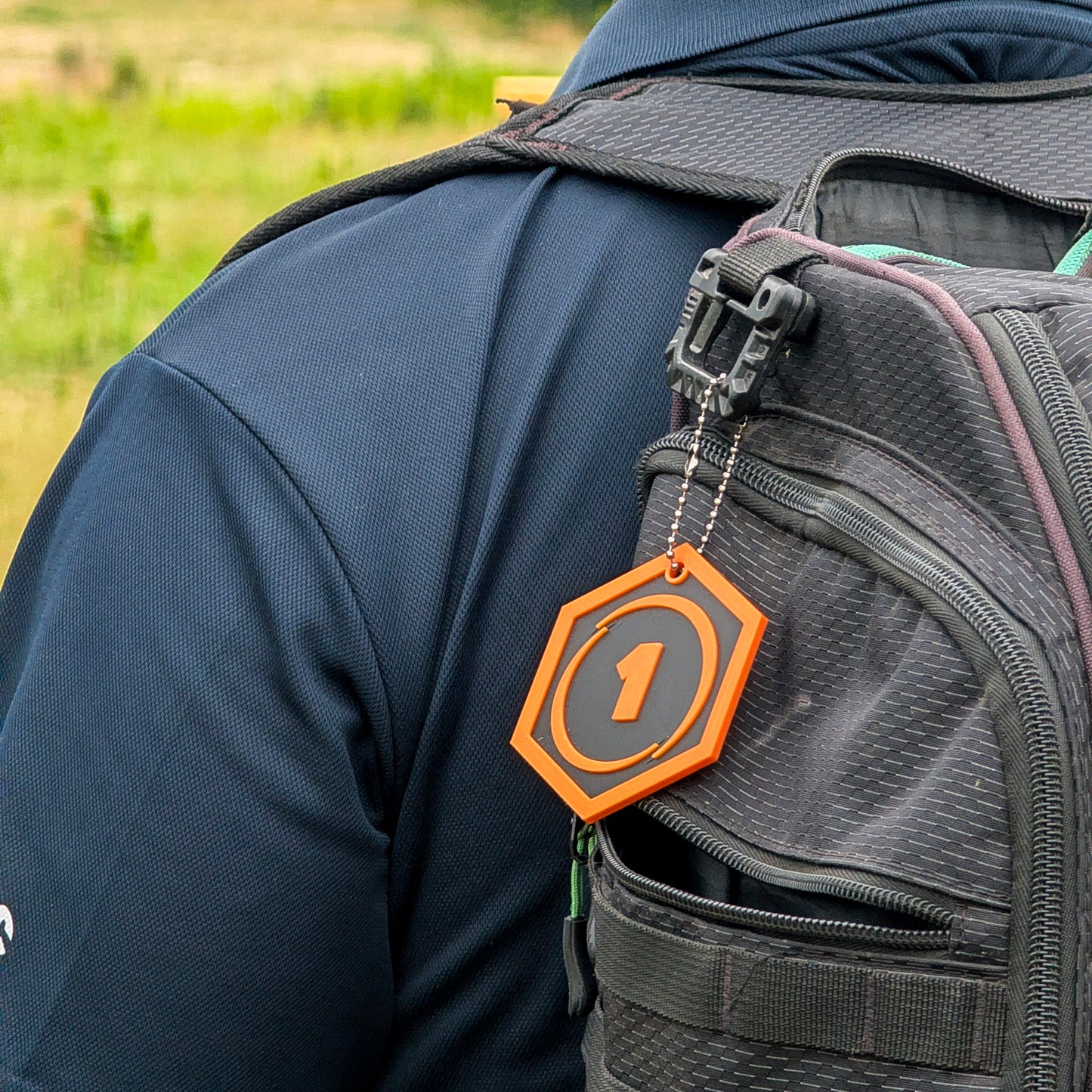 Close-up of a person wearing a dark navy blue shirt and carrying a black backpack outdoors. A bright orange hexagonal disc golf bag tag