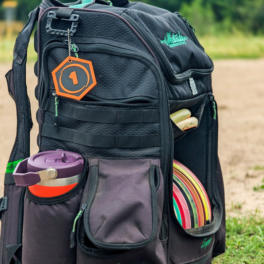 A black disc golf backpack with multiple pockets, shown outdoors on a dirt path with grass in the background. The backpack features a purple water bottle in a side pocket, a stack of colorful disc golf discs, and a green logo on the upper right. And a minimates bag tag hanging from the side